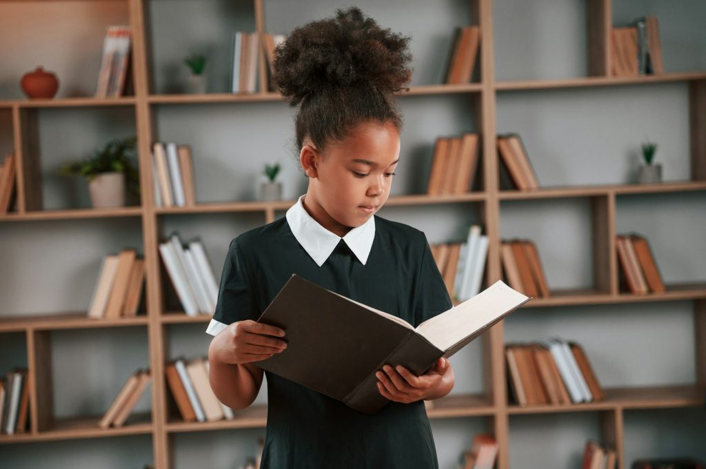 Holding book and reading. Cute african american girl in school uniform is at home library